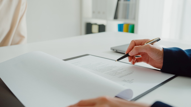 Image of a contract being reviewed. A suited person points to it with a pen, suggesting attention to detail and careful consideration, for use in content creation.